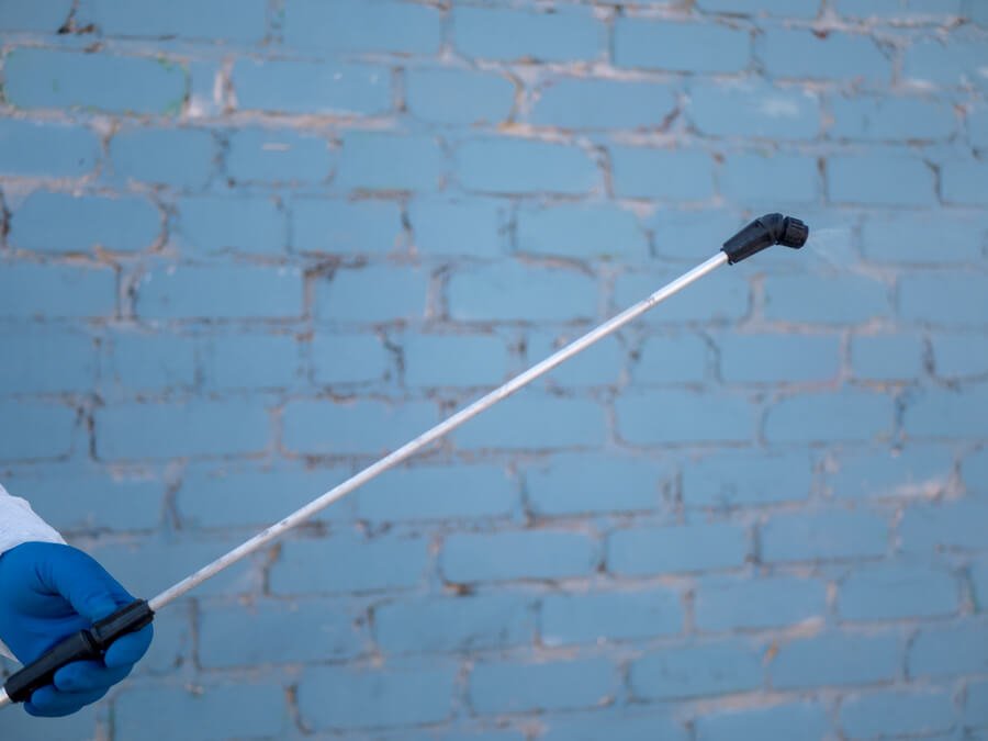 Gloved hand holding a pest control spray wand in front of a blue brick wall.
