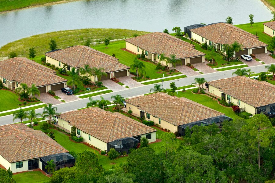 Aerial view of suburban homes with tiled roofs, palm trees, and a lake in the background.

