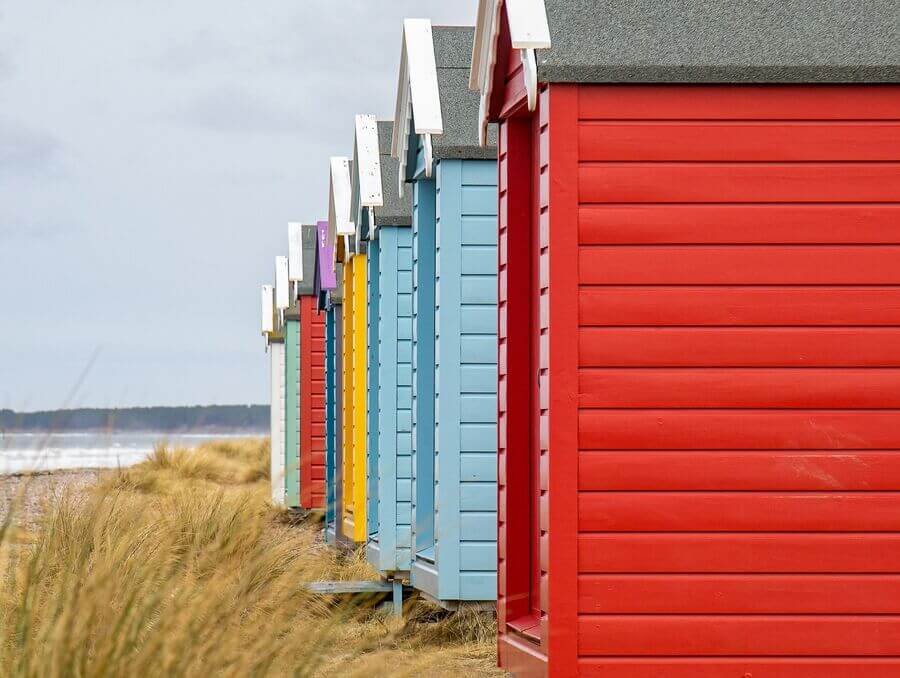 Colorful beach huts lined up along a sandy shoreline with tall grass.