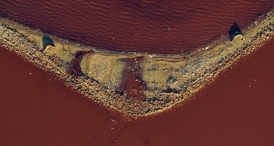 Aerial view of reddish water meeting a sandy shoreline, forming a sharp V-shaped coastline with scattered rocks and people visible.