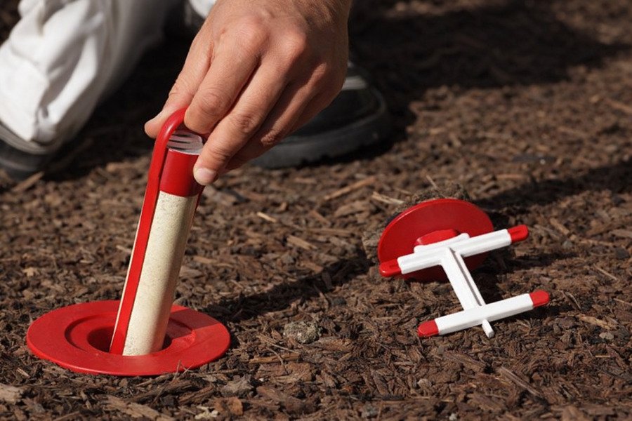 Fast termite treatment - Hand placing a red termite bait station into soil with tool beside it.